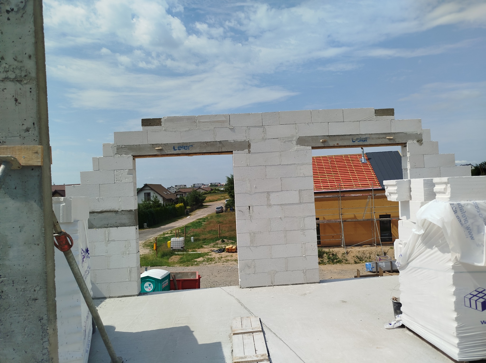 Wznoszenie muru z białych bloczków, widok przez otwór okienny na dom z czerwoną dachówką i błękitne niebo. Częściowo widoczne materiały budowlane i podpory.