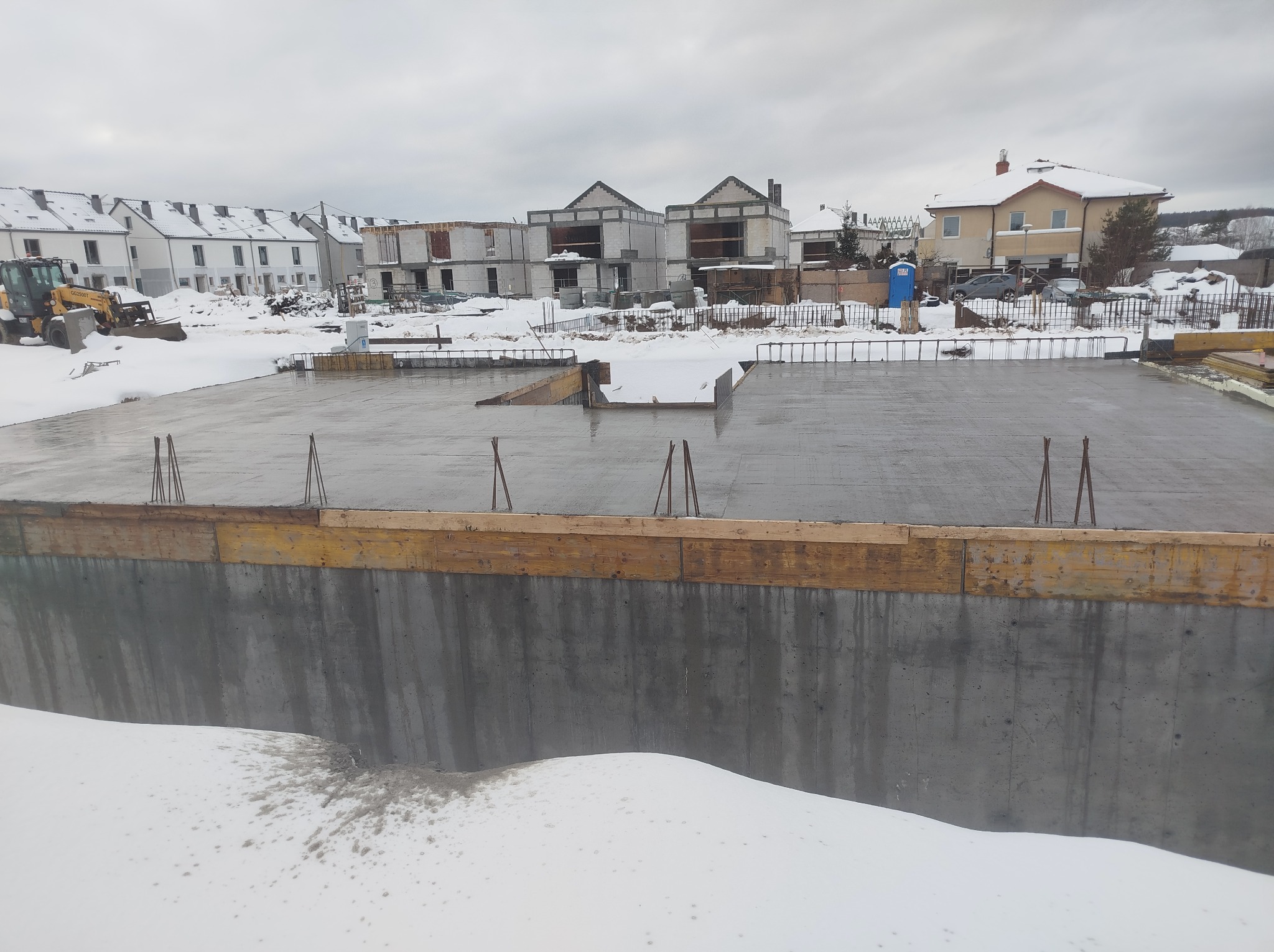 Zalany fundament pod budowę domu jednorodzinnego w zimowej scenerii. Widoczne szalunki i wystające pręty zbrojeniowe. W tle zabudowa osiedlowa i pochmurne niebo.