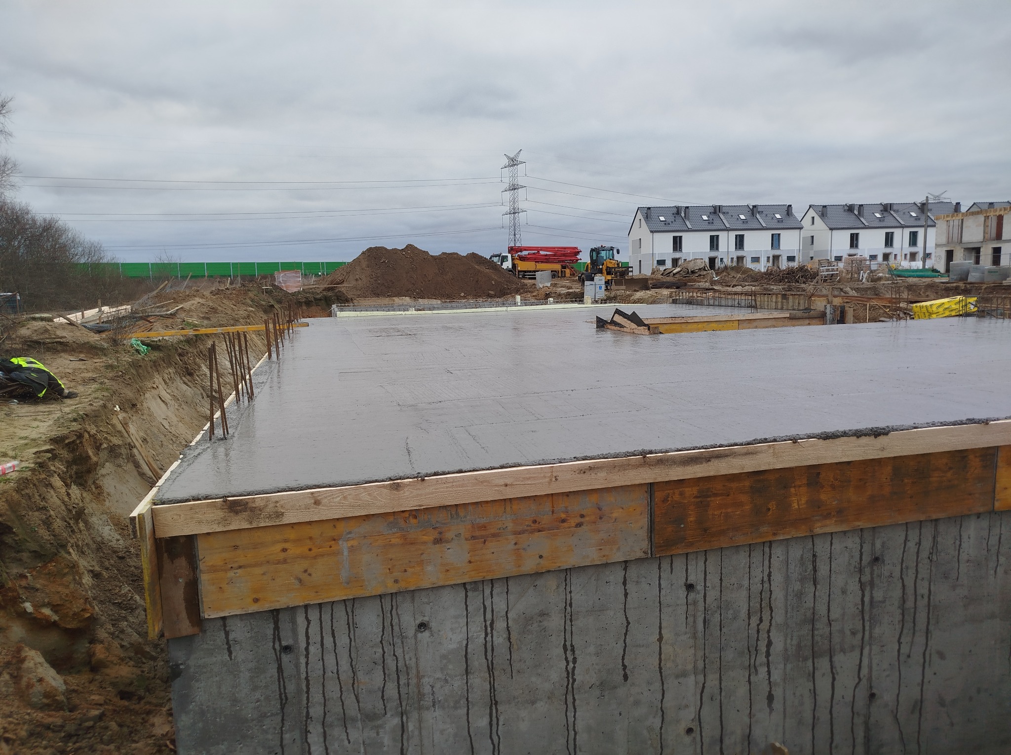 Zalany beton na fundamencie z drewnianym szalunkiem, widok z bliska na betonową konstrukcję. W tle budowa osiedla i sprzęt budowlany.