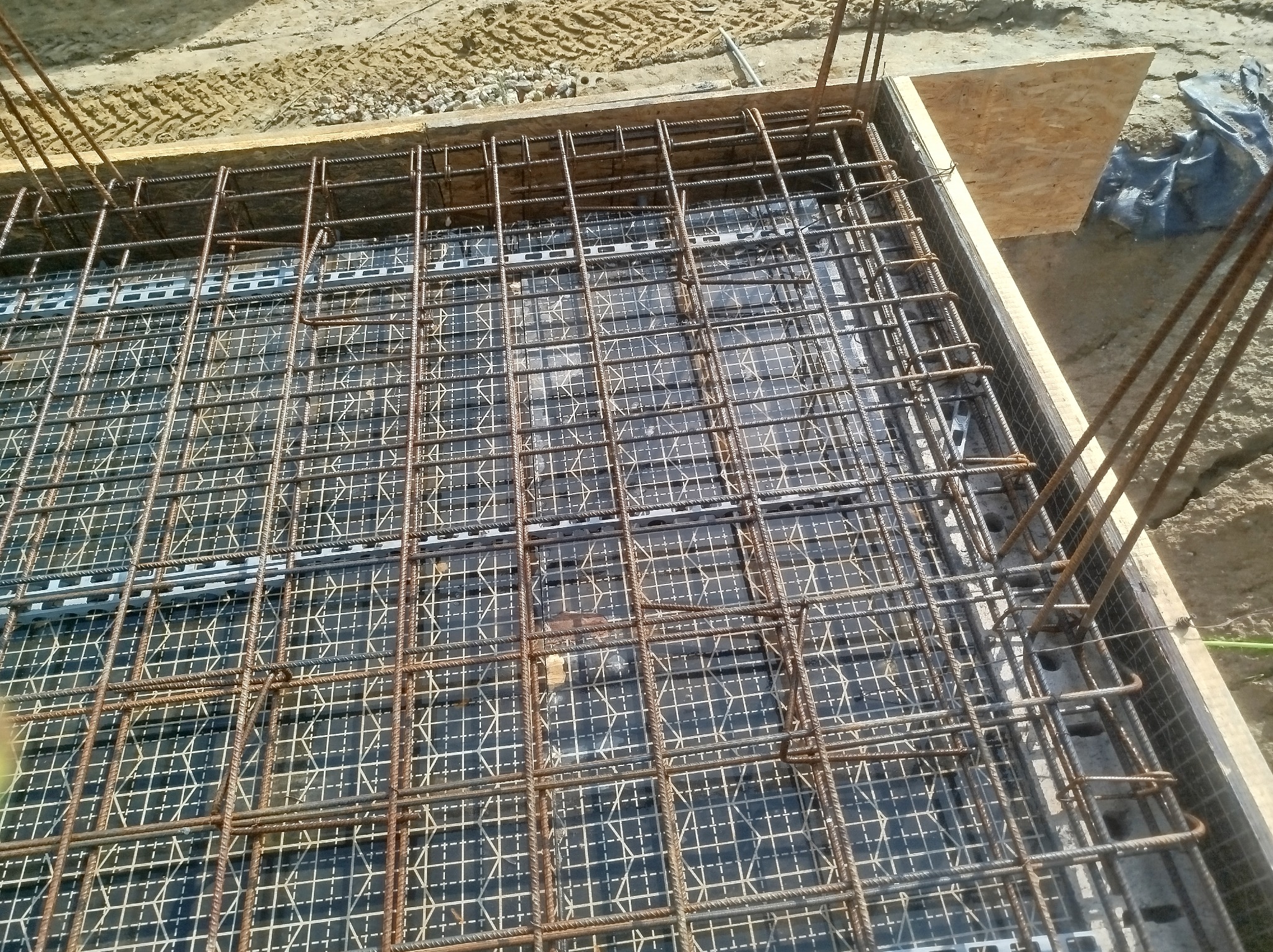 Zbrojenie fundamentów w szalunku z widoczną siatką zbrojeniową i wystającymi prętami pionowymi. Widok z góry na przygotowany fundament pod budowę.