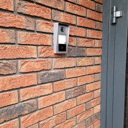 TimeOut System - Szary wideodomofon IP zamontowany na czerwono-ceglanej ścianie obok szarej ramy drzwiowej.