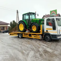 Żółto-zielony traktor John Deere 6320 zabezpieczony pasami na żółtej platformie pojazdu pomocy drogowej Iveco Eurocargo z widocznym logo firmy Sapinski na dachu, w tle budynek i zaśnieżony teren.