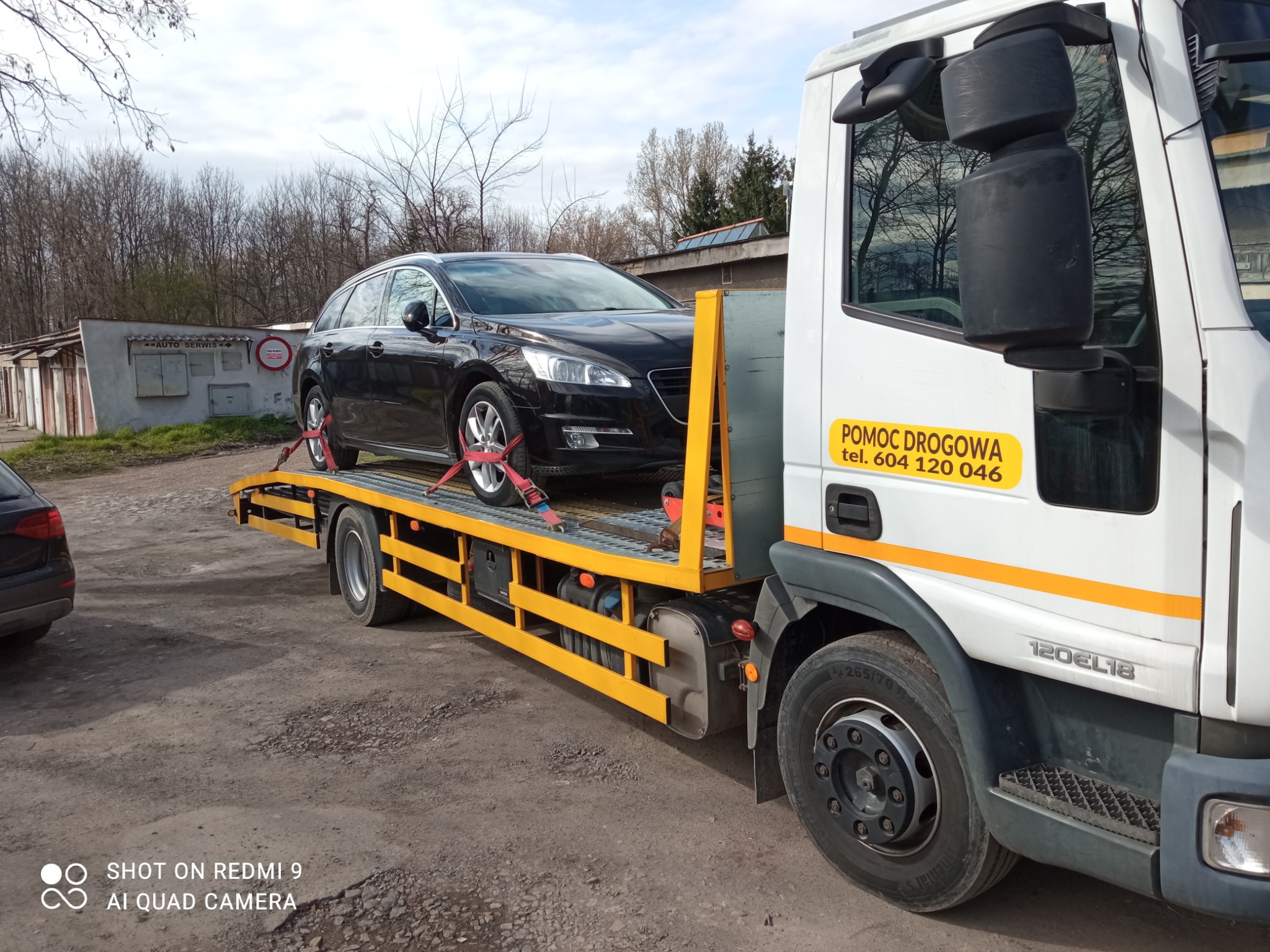 Czarny samochód osobowy Peugeot na żółtej platformie lawety pomocy drogowej z naklejką 'POMOC DROGOWA tel. 604 120 046' na kabinie, w tle budynki garażowe i drzewa bez liści.