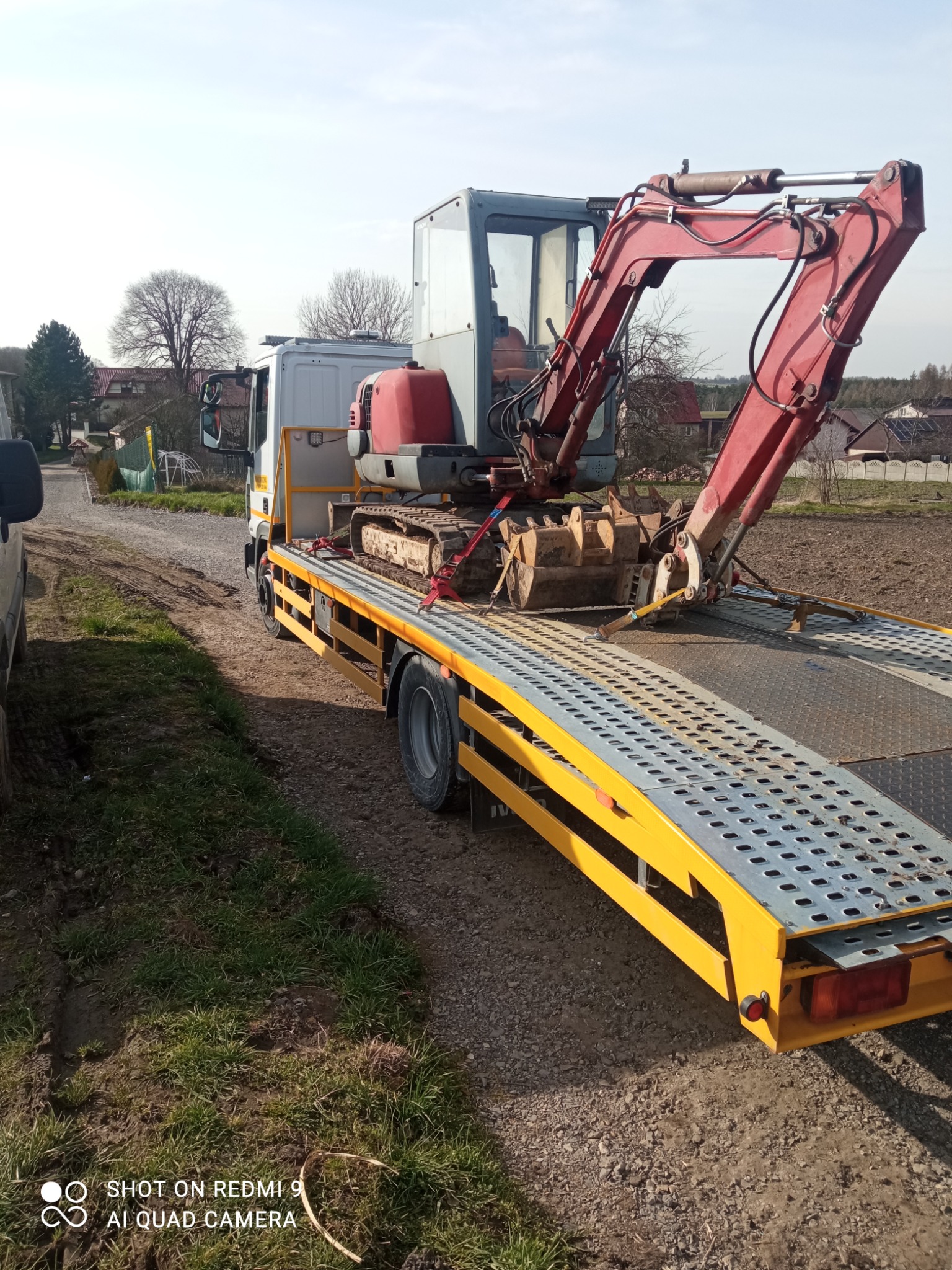 Żółta platforma transportowa przewożąca małą czerwoną koparkę na tle wiejskiego krajobrazu z polami i zabudowaniami.