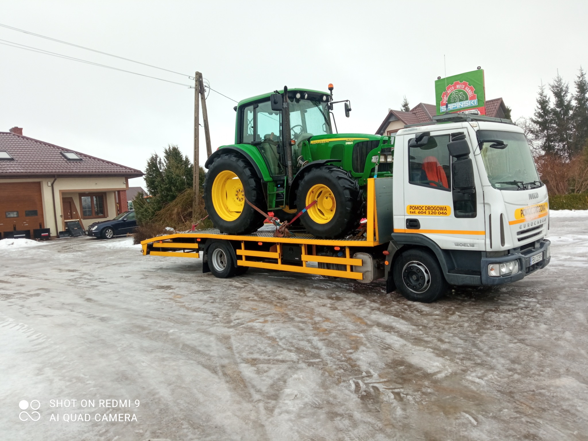 Żółto-zielony traktor John Deere 6320 zabezpieczony pasami na żółtej platformie pojazdu pomocy drogowej Iveco Eurocargo z widocznym logo firmy Sapinski na dachu, w tle budynek i zaśnieżony teren.