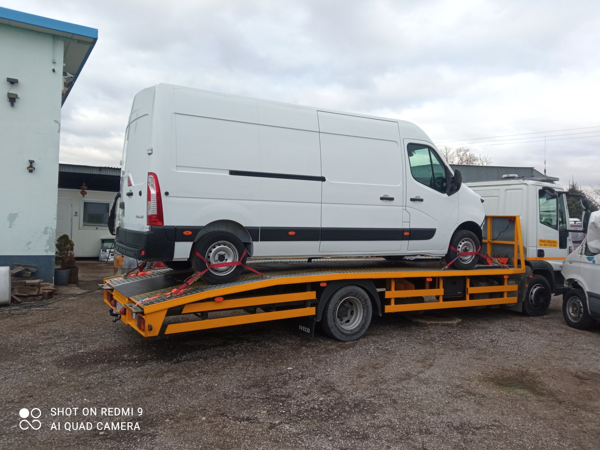 Biały furgon Renault Master zabezpieczony czerwonymi pasami transportowymi na żółtej platformie pojazdu pomocy drogowej Iveco, w tle budynek i pochmurne niebo.