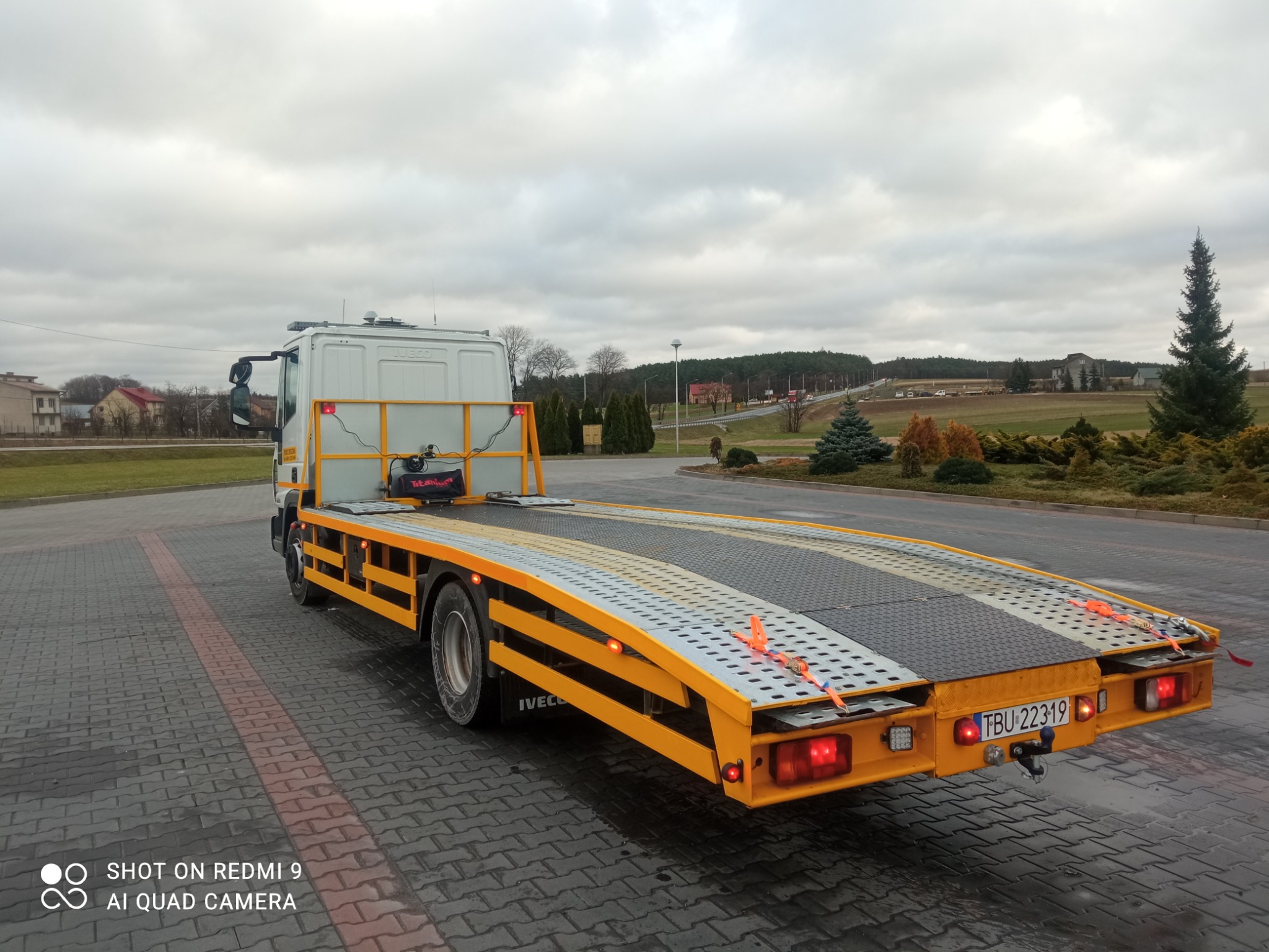 Pojazd specjalny z żółto-czarną platformą do transportu pojazdów, zaparkowany na brukowanej powierzchni, widok z tyłu, na tablicy rejestracyjnej widoczny numer TBU 22319.