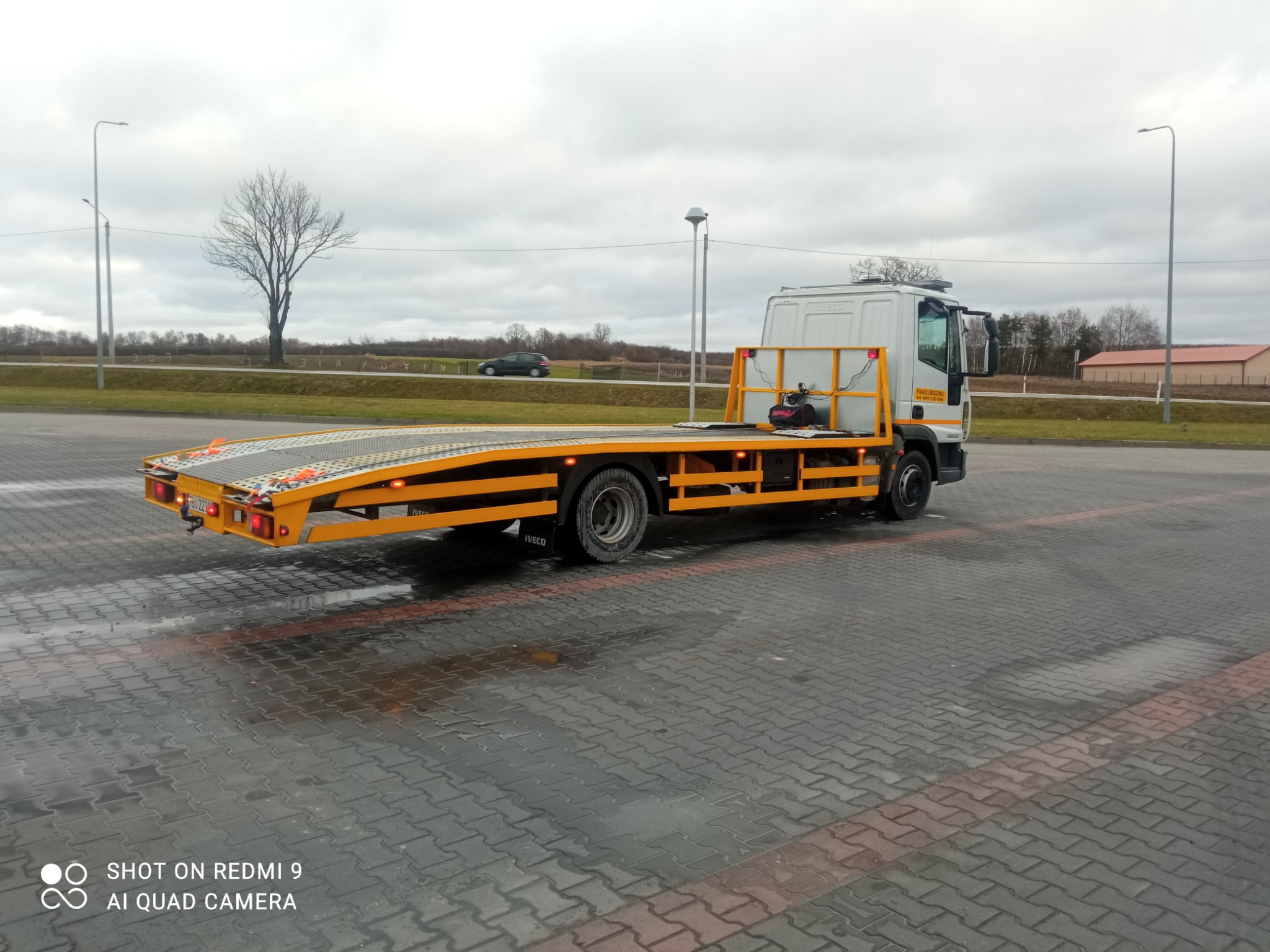 Żółto-biała ciężarówka z platformą na parkingu z mokrą kostką brukową, widoczne budynki w tle i pochmurne niebo.