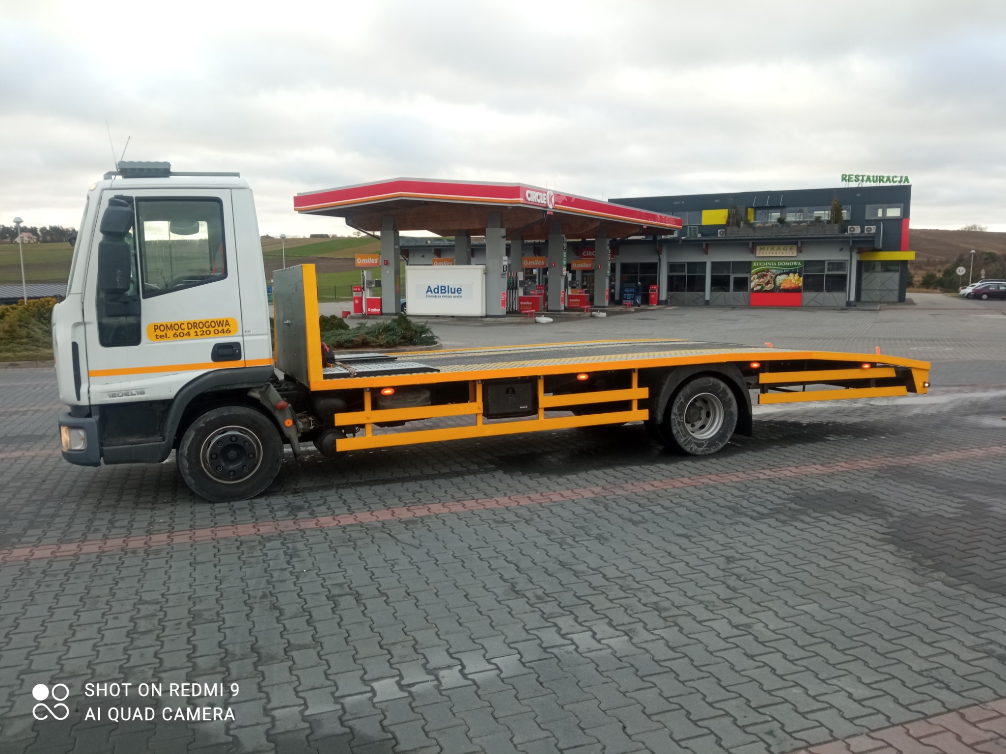 Żółta ciężarówka z platformą do transportu pojazdów, zaparkowana na kostce brukowej przed stacją benzynową Circle K i restauracją Mirage.