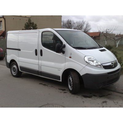 Biały Opel Vivaro z przesuwanymi drzwiami bocznymi, zaparkowany na szarej nawierzchni, widok z boku na tle budynków i drzew.