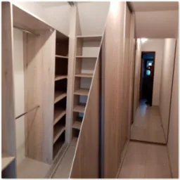 Nowoczesna garderoba z przesuwnymi drzwiami i półkami w kolorze jasnego drewna, widok na korytarz z lustrem i odbijającą się sylwetką fotografa.