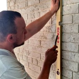 Decor Home - Mężczyzna przykłada poziomicę do ściany z białej cegły, trzymając w dłoni nóż do cięcia.