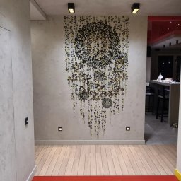 Decor Home - Ściana w przedpokoju pokryta dekoracyjnym mikrocementem z wzorem mandali w odcieniach czerni i złota, oświetlona punktowymi lampami sufitowymi i ściennymi, z widoczną podłogą z jasnego drewna.