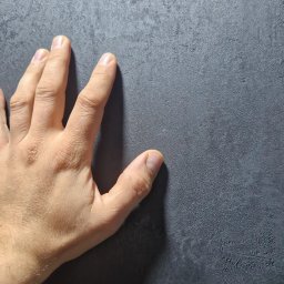 Decor Home - Dłoń dotyka szarej, chropowatej powierzchni ściany z efektem betonu, widoczne nieregularne wzory i faktura.