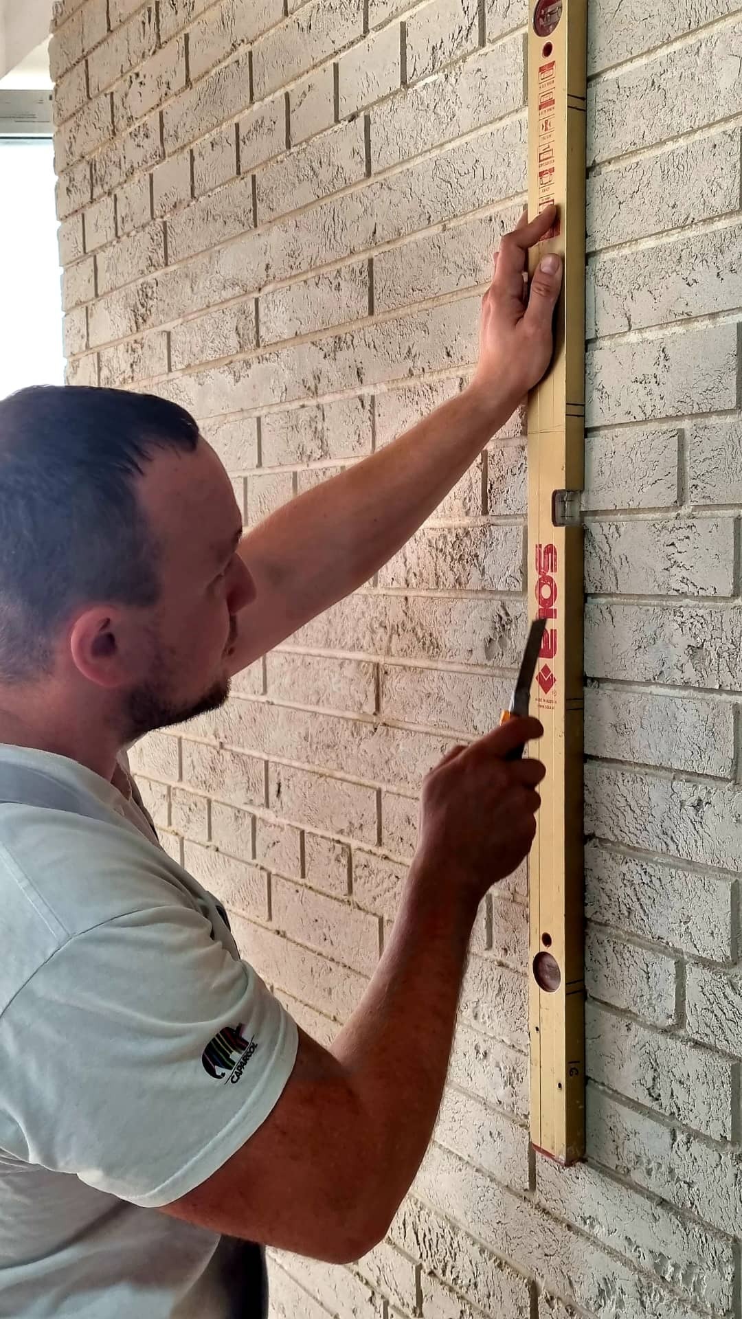 Mężczyzna przykłada poziomicę do ściany z białej cegły, trzymając w dłoni nóż do cięcia.