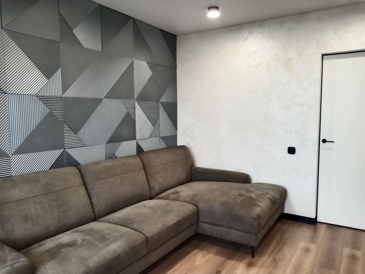 Szara sofa narożna na drewnianej podłodze, ściana z geometrycznym wzorem 3D oraz biała ściana z efektem dekoracyjnym, czarne detale wykończeniowe.