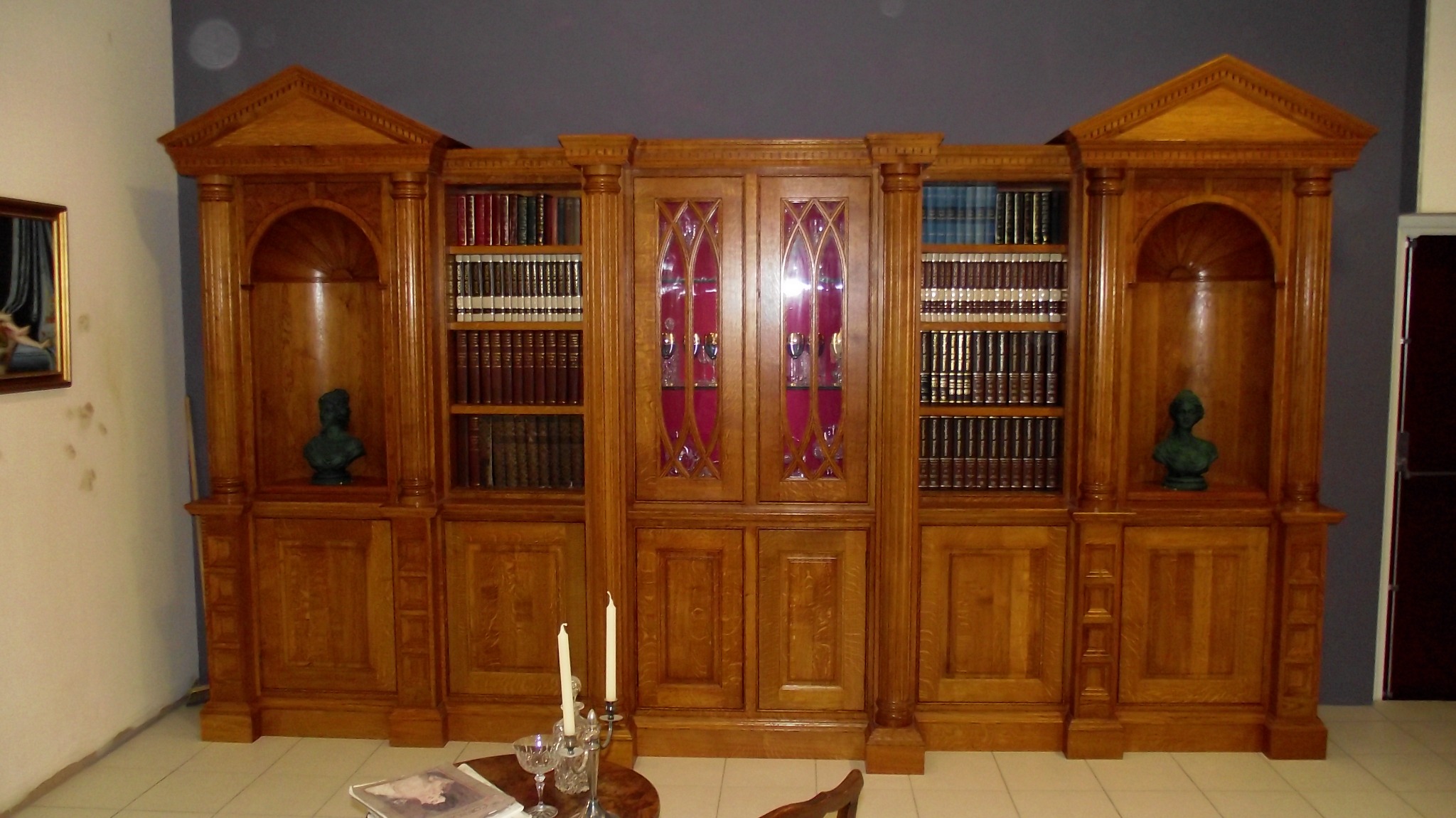 Klasyczna, drewniana biblioteczka z przeszklonymi drzwiczkami, kolumnami i wnękami z rzeźbami, wypełniona książkami, oświetlona świecami na stoliku.