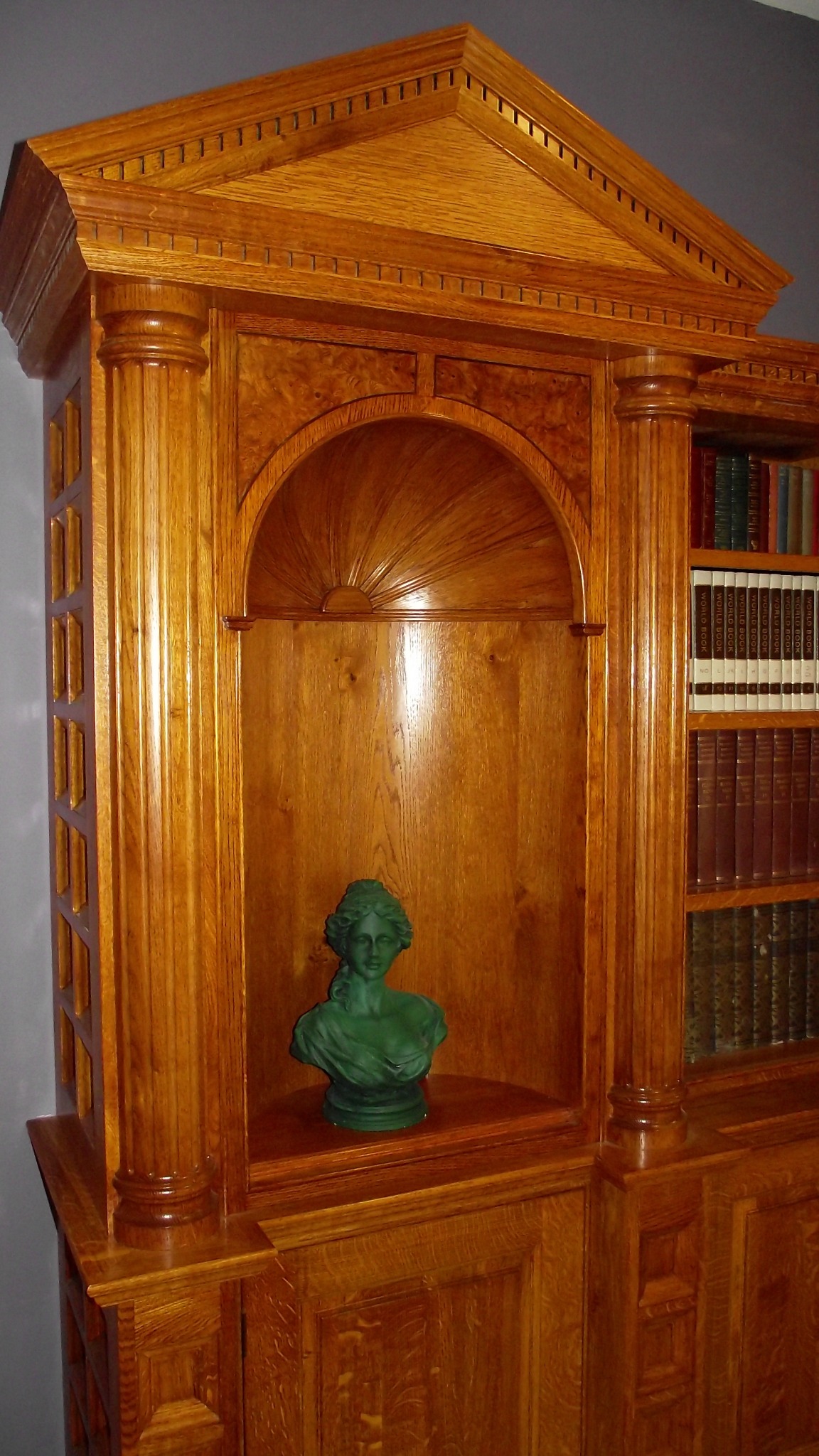 Klasyczna, dębowa biblioteczka z kolumnami i niszą z rzeźbą, wypełniona książkami.