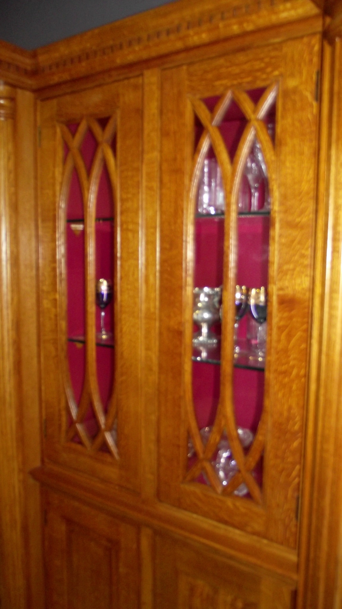 Drewniana szafa wnękowa z przeszkleniami w kształcie gotyckich łuków, podświetlona czerwonym tłem, eksponująca kieliszki i puchary na szklanych półkach.