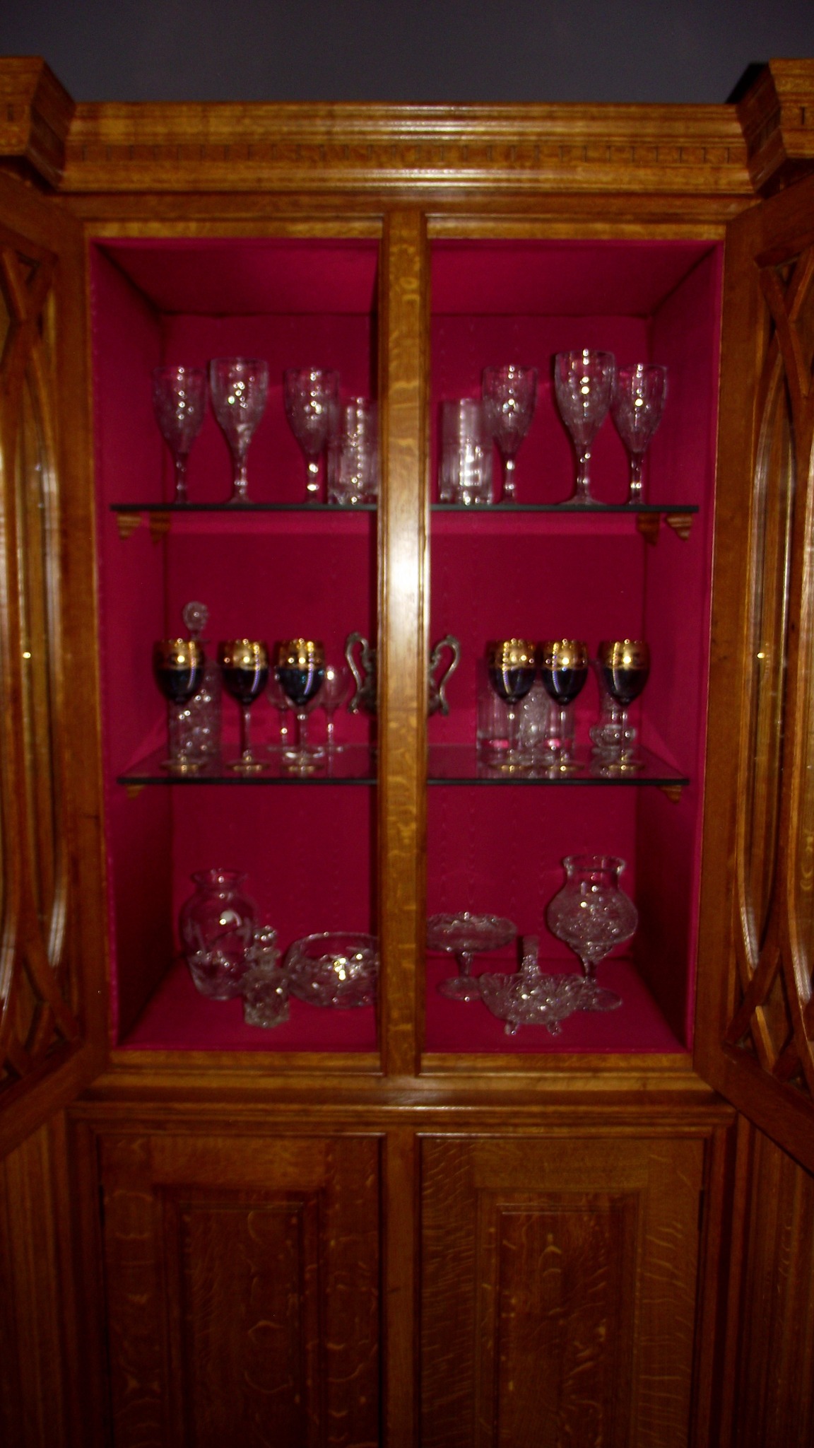 Otwarta, drewniana witryna z czerwonym aksamitnym wnętrzem, wypełniona kryształowymi kieliszkami i misami na szklanych półkach.
