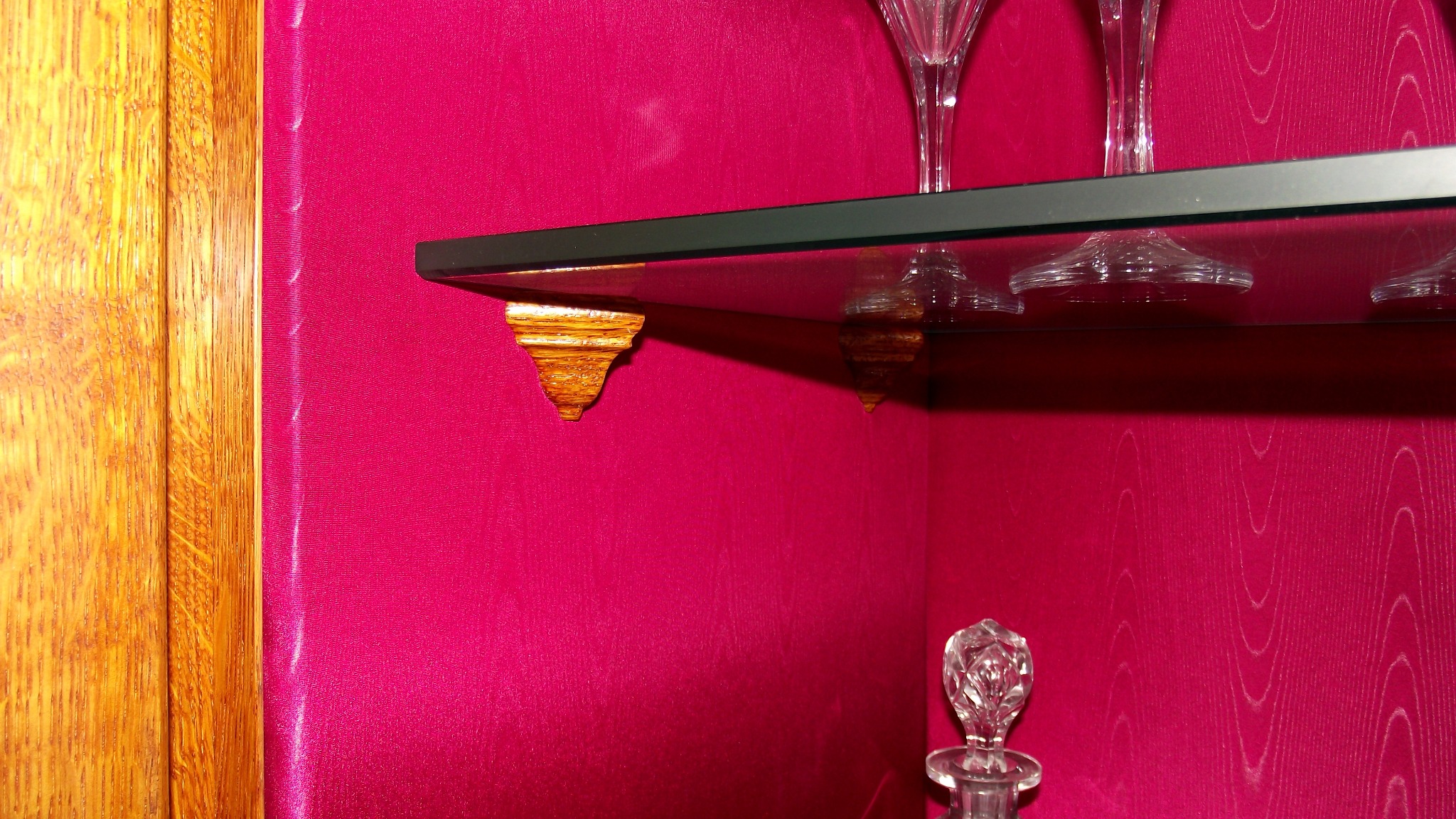 Szklana półka wewnątrz szafy z różowym wykończeniem, podparta ozdobnymi, złotymi wspornikami, na półce kieliszki i kryształowa karafka.