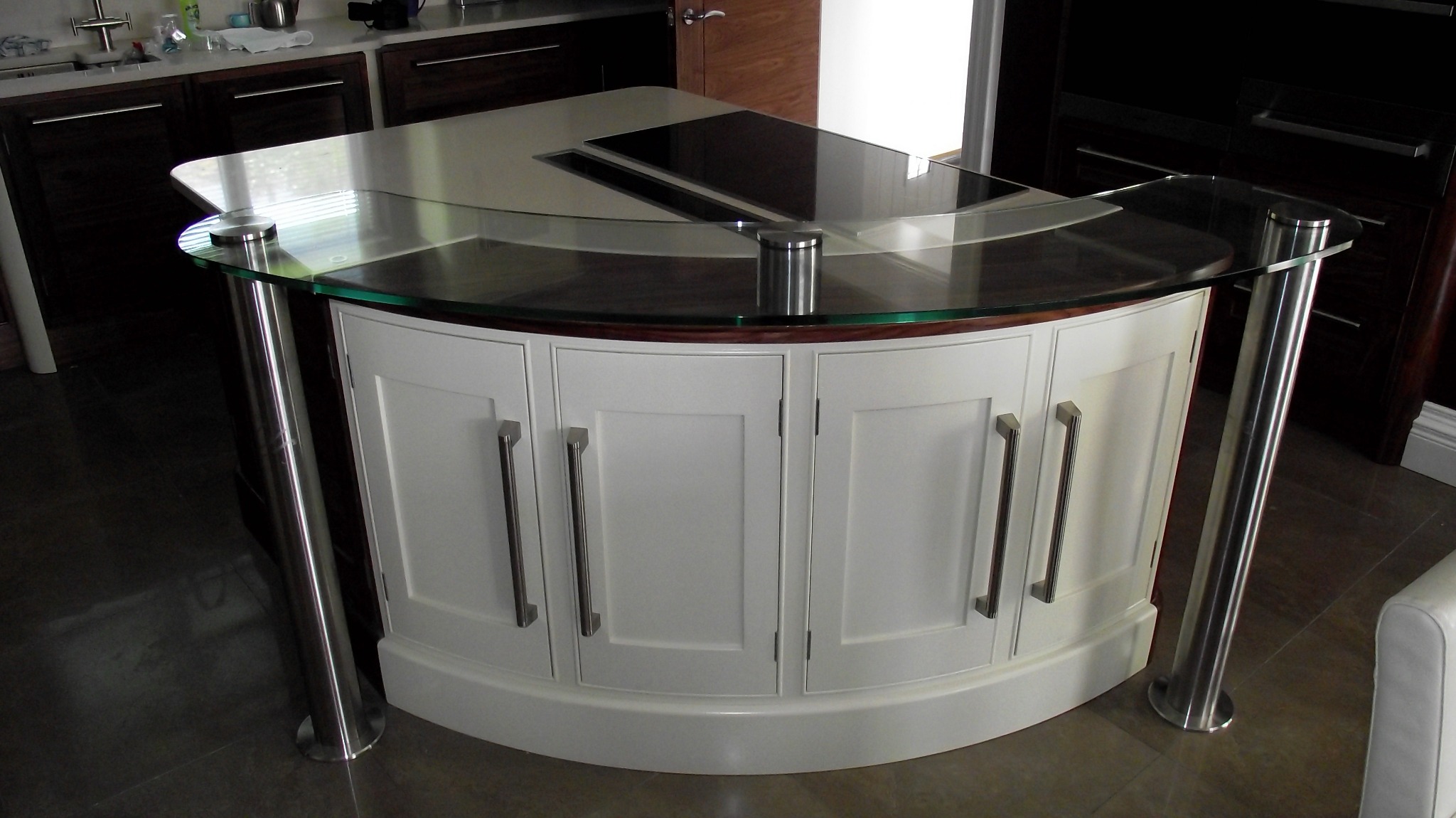Półokrągła wyspa kuchenna z białymi frontami szafek, blatem z ciemnego drewna i szklaną nadstawką, wsparta na metalowych nogach. W blacie zamontowana płyta indukcyjna.