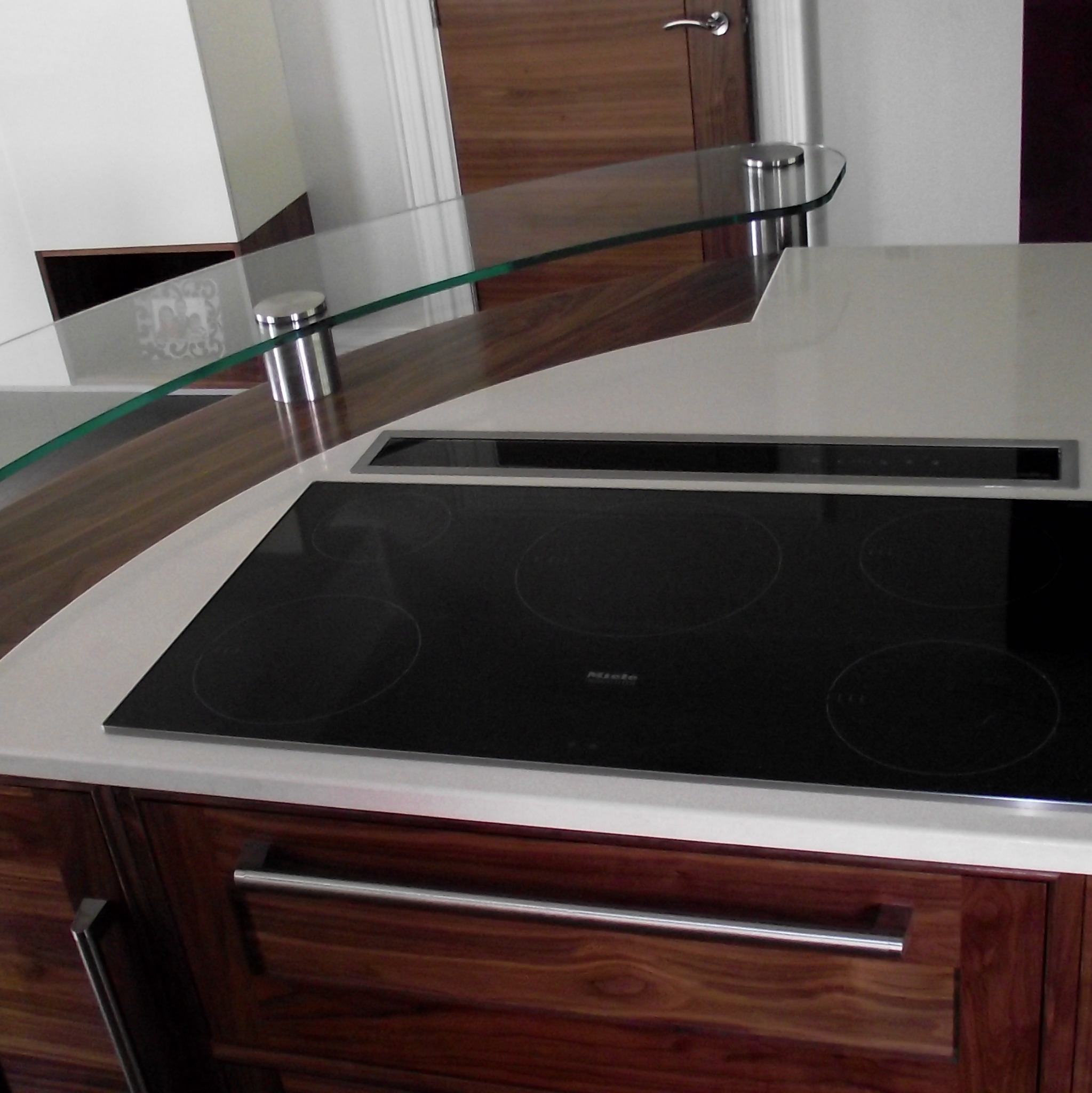 Nowoczesna kuchnia z drewnianymi szafkami, białym blatem, czarną płytą indukcyjną Miele i szklaną półką na metalowych wspornikach