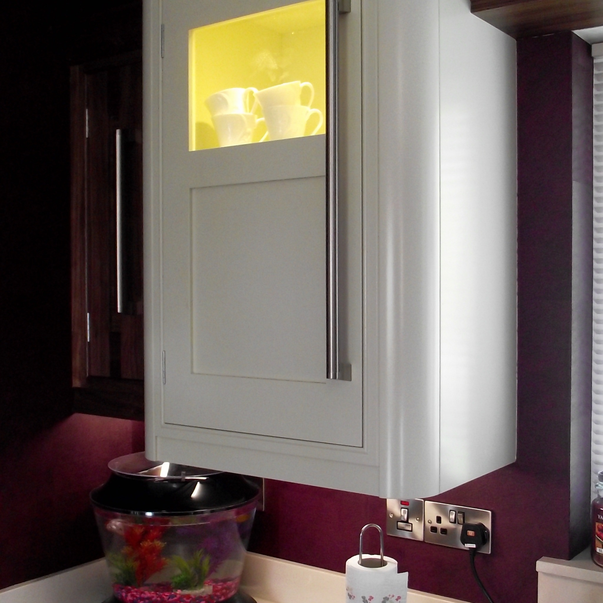 Biała szafka kuchenna z podświetlaną witryną, w której stoją białe filiżanki, zamontowana nad blatem, obok ciemnobrązowej szafki i bordowej ściany z gniazdkami.