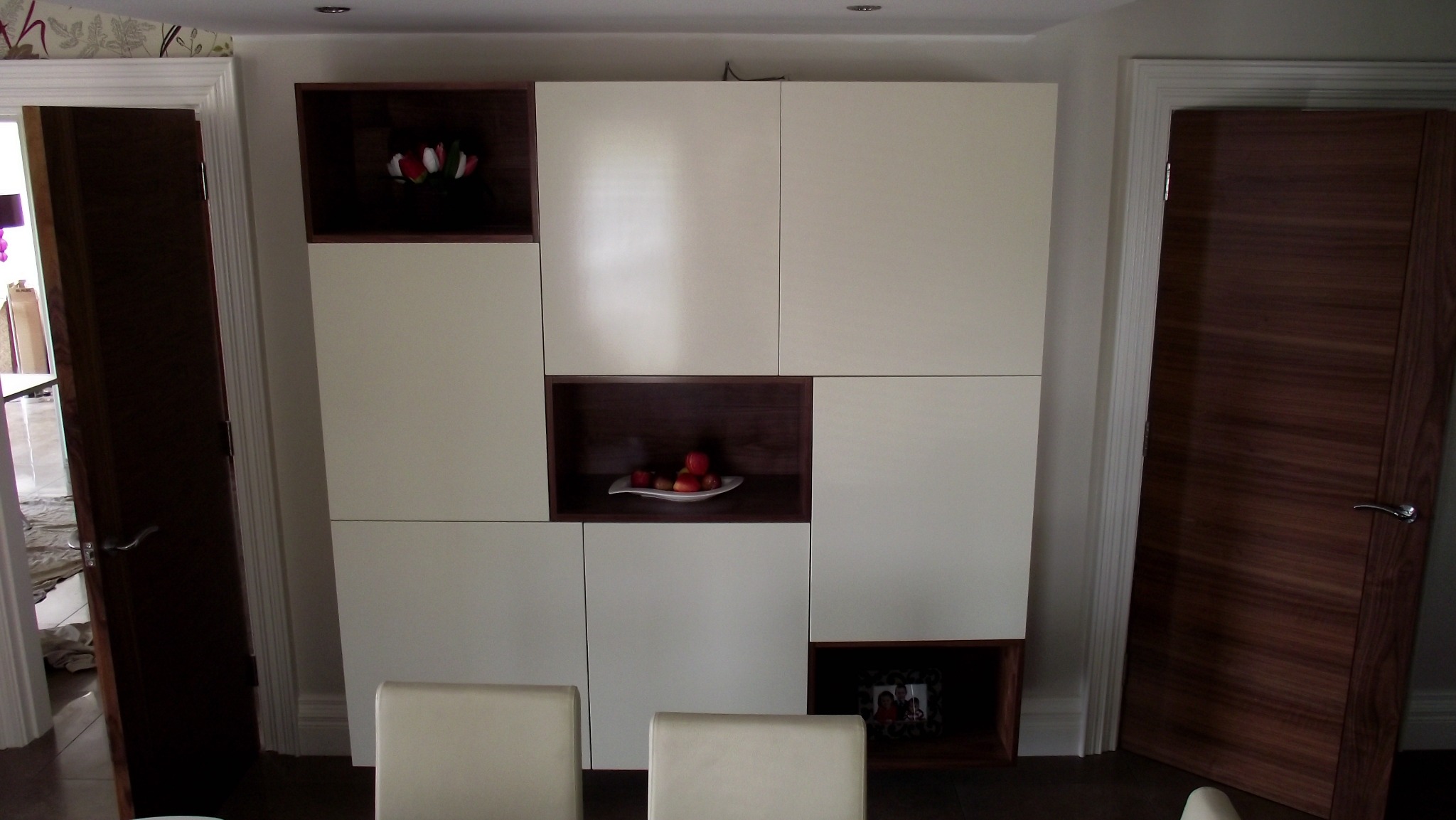 Biała szafa wnękowa z ciemnymi wstawkami, z otwartymi półkami z dekoracjami: kwiatami, owocami i ramką ze zdjęciem, umieszczona między drzwiami.