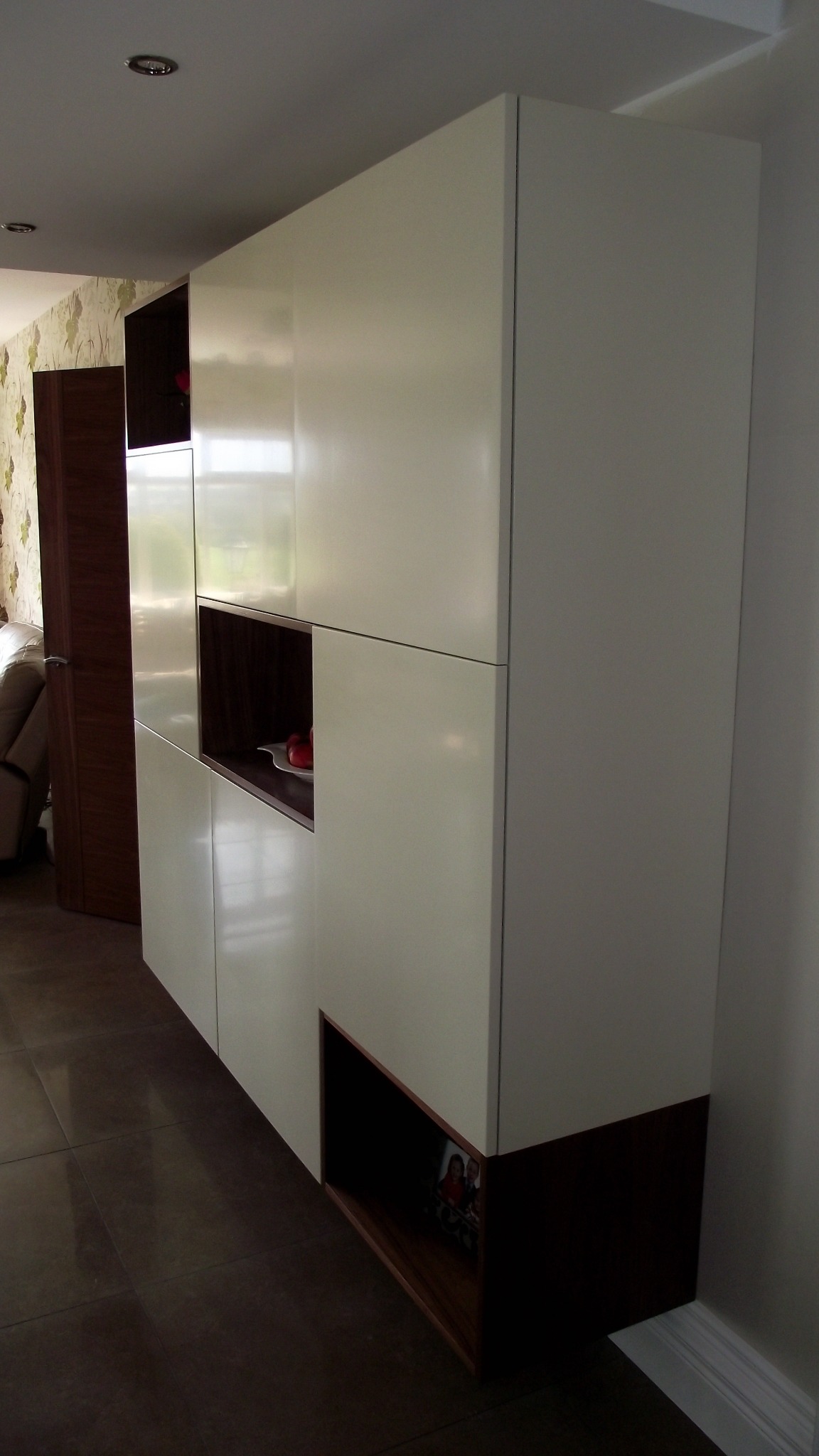 Nowoczesna biało-brązowa zabudowa meblowa z otwartymi półkami, na jednej z nich dekoracyjne jabłka, na dolnej ramka ze zdjęciem, widoczna na tle brązowej podłogi.