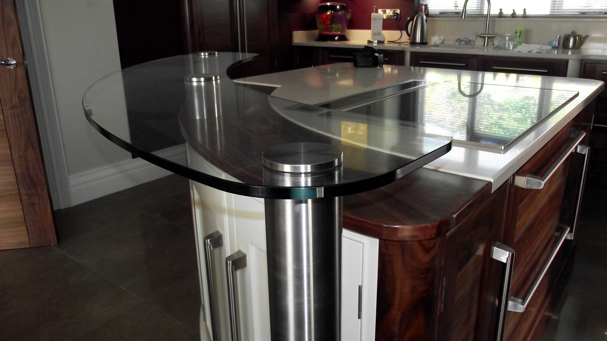 Szklany blat zaokrąglony na wyspie kuchennej, wsparty na metalowych cylindrycznych nogach, z widocznym fragmentem blatu ze stali nierdzewnej i drewnianych szafek w tle.