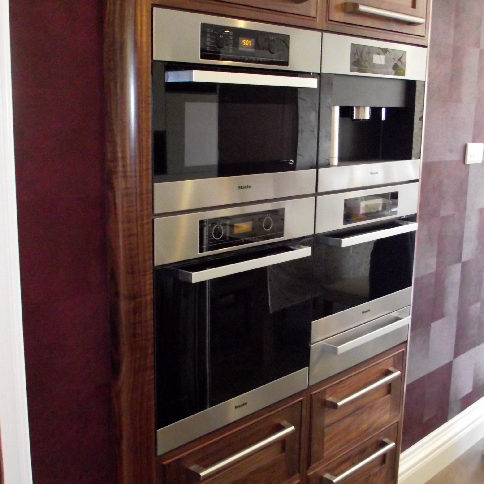 Zabudowa kuchenna z ciemnego drewna z wbudowanymi urządzeniami Miele: piekarnik, mikrofalówka i ekspres do kawy, na tle ściany z tapetą w geometryczny wzór.