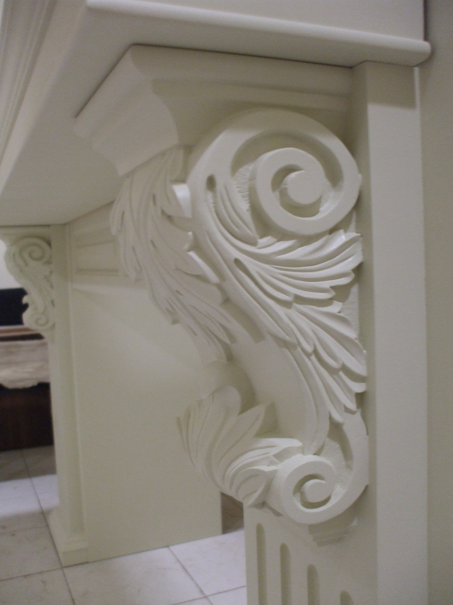 Biały, bogato zdobiony element architektoniczny z motywem roślinnym i spiralnym, prawdopodobnie fragment portalu lub obudowy kominka, widziany z bliska.