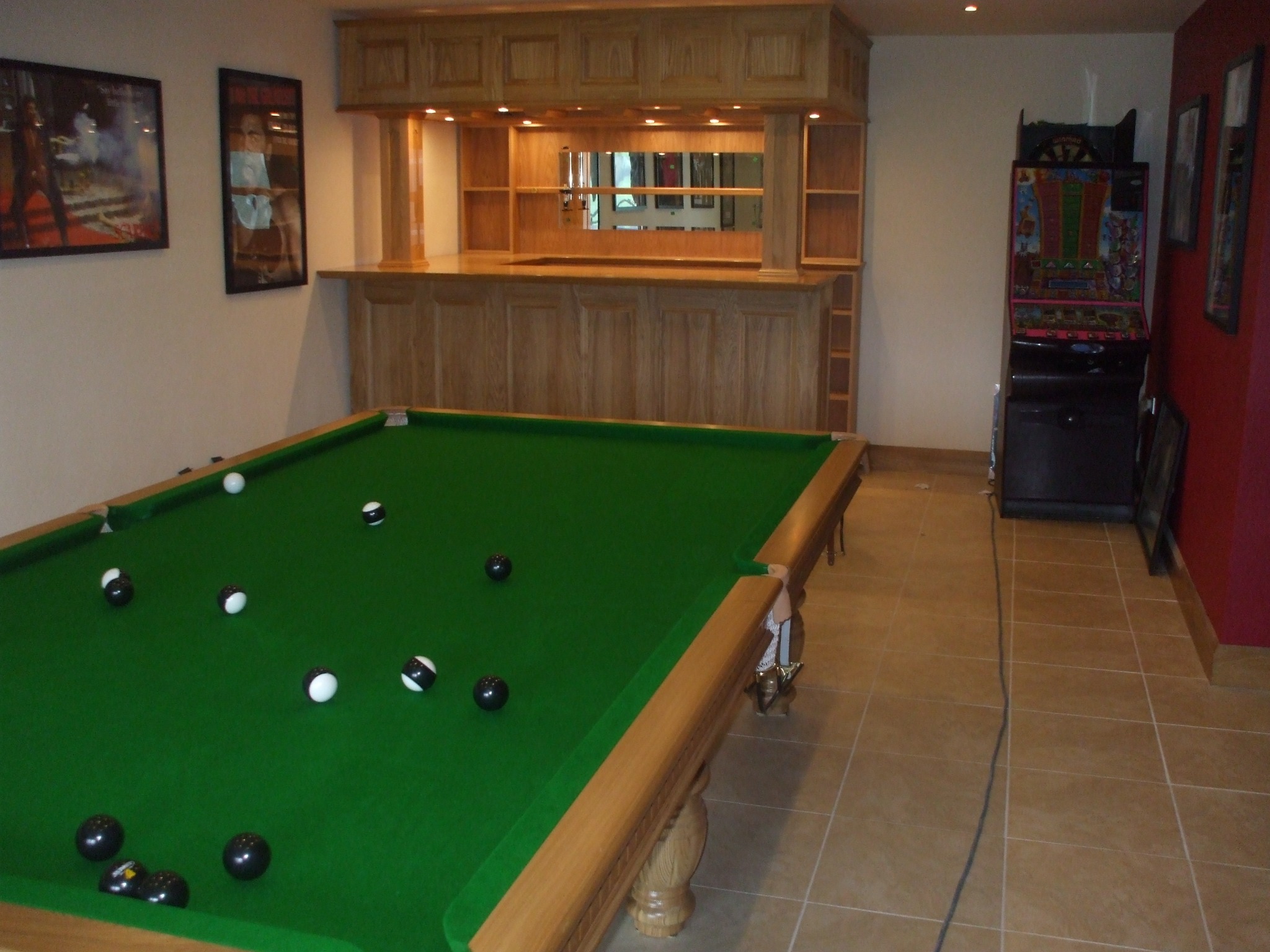 Pokój rekreacyjny z drewnianym barem, stołem bilardowym z rozrzuconymi bilami i automatem do gier w tle.