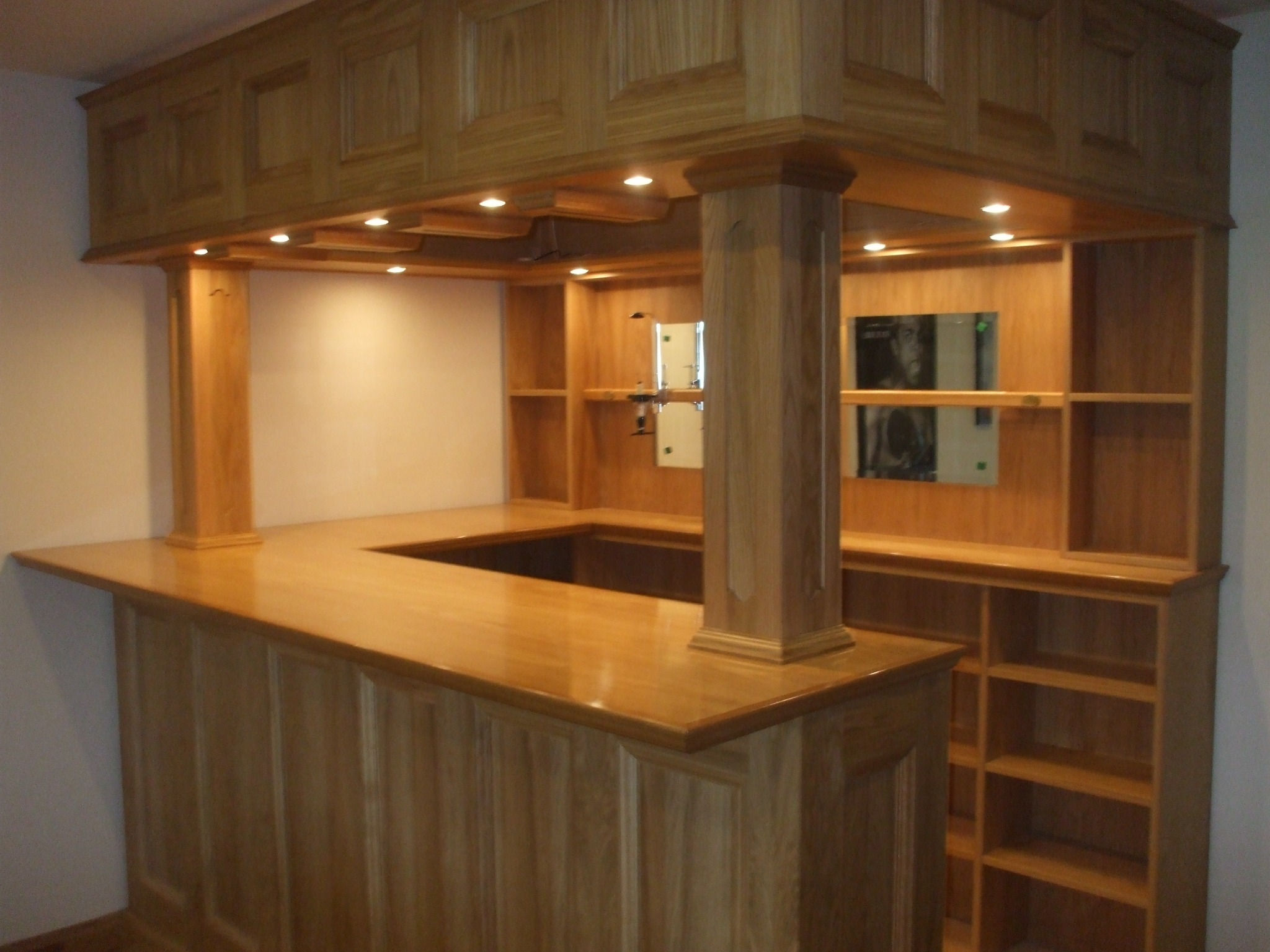 Drewniany bar z podświetleniem LED, blatem z wyciętym otworem, kolumną dekoracyjną i półkami w tle.