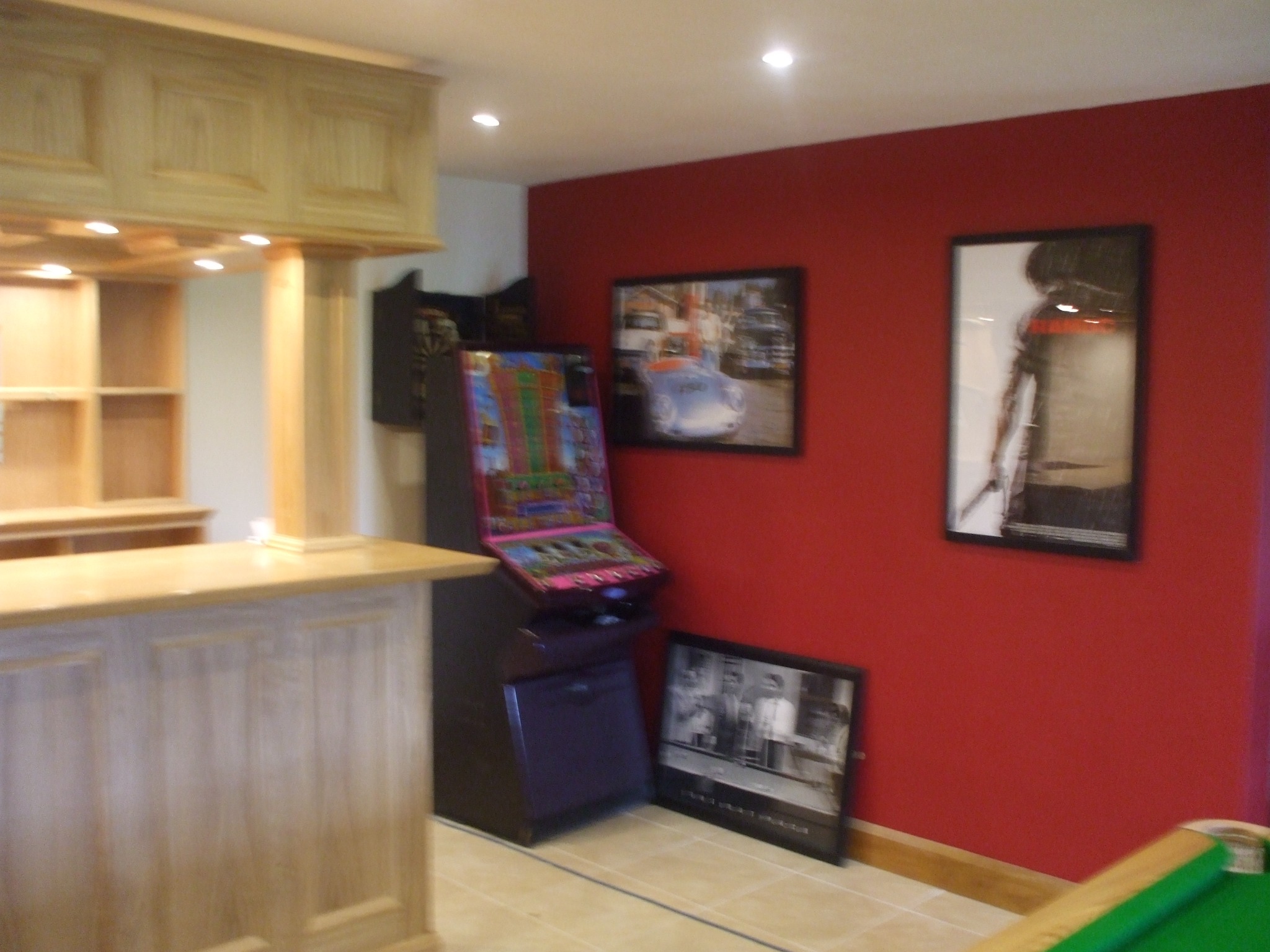 Drewniany bar z oświetleniem i półkami w pomieszczeniu z czerwonymi ścianami, automat do gier i obrazy na ścianie.