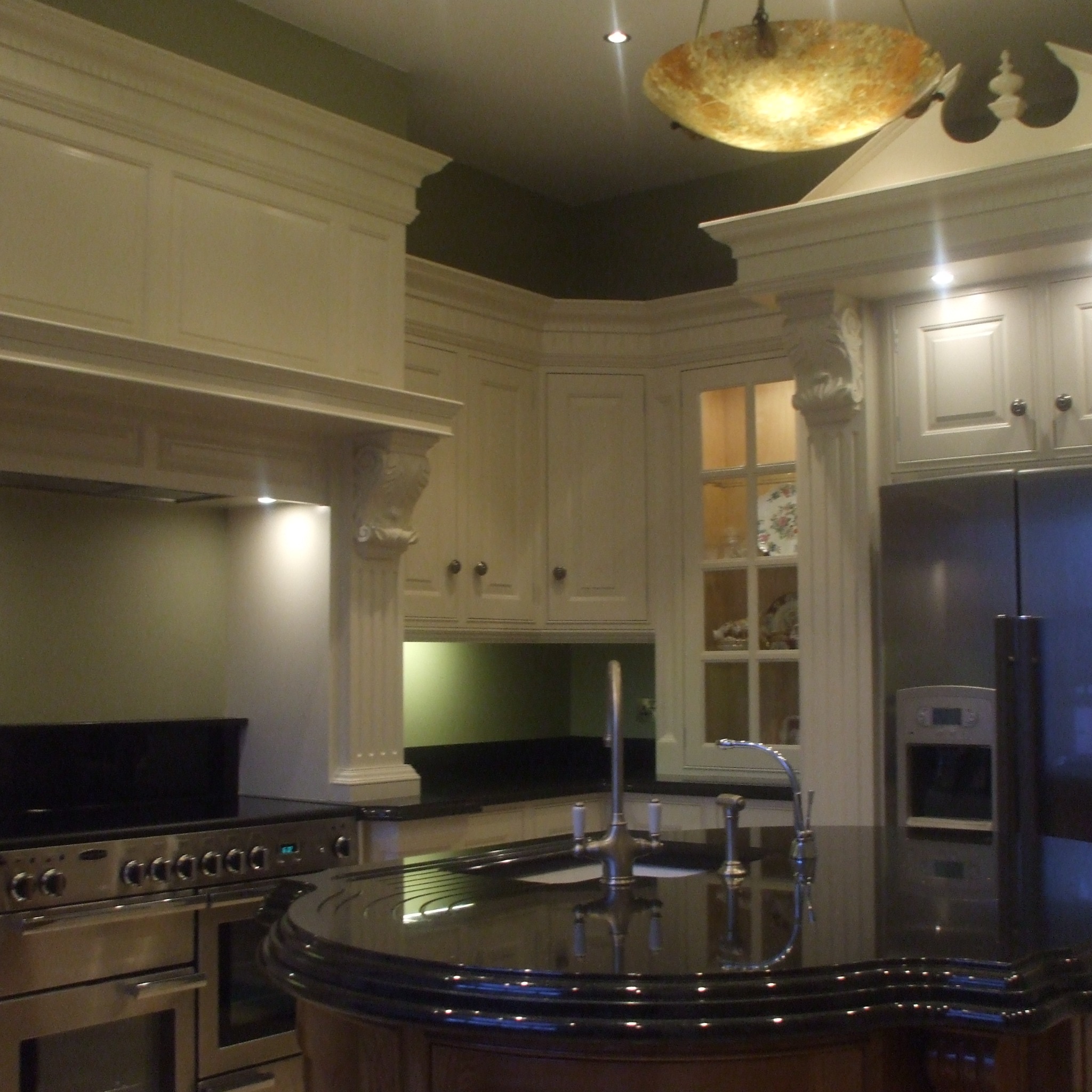 Elegancka kuchnia z wyspą i blatem z czarnego kamienia, ozdobnymi kolumnami i okapem, oświetlona dekoracyjną lampą sufitową.