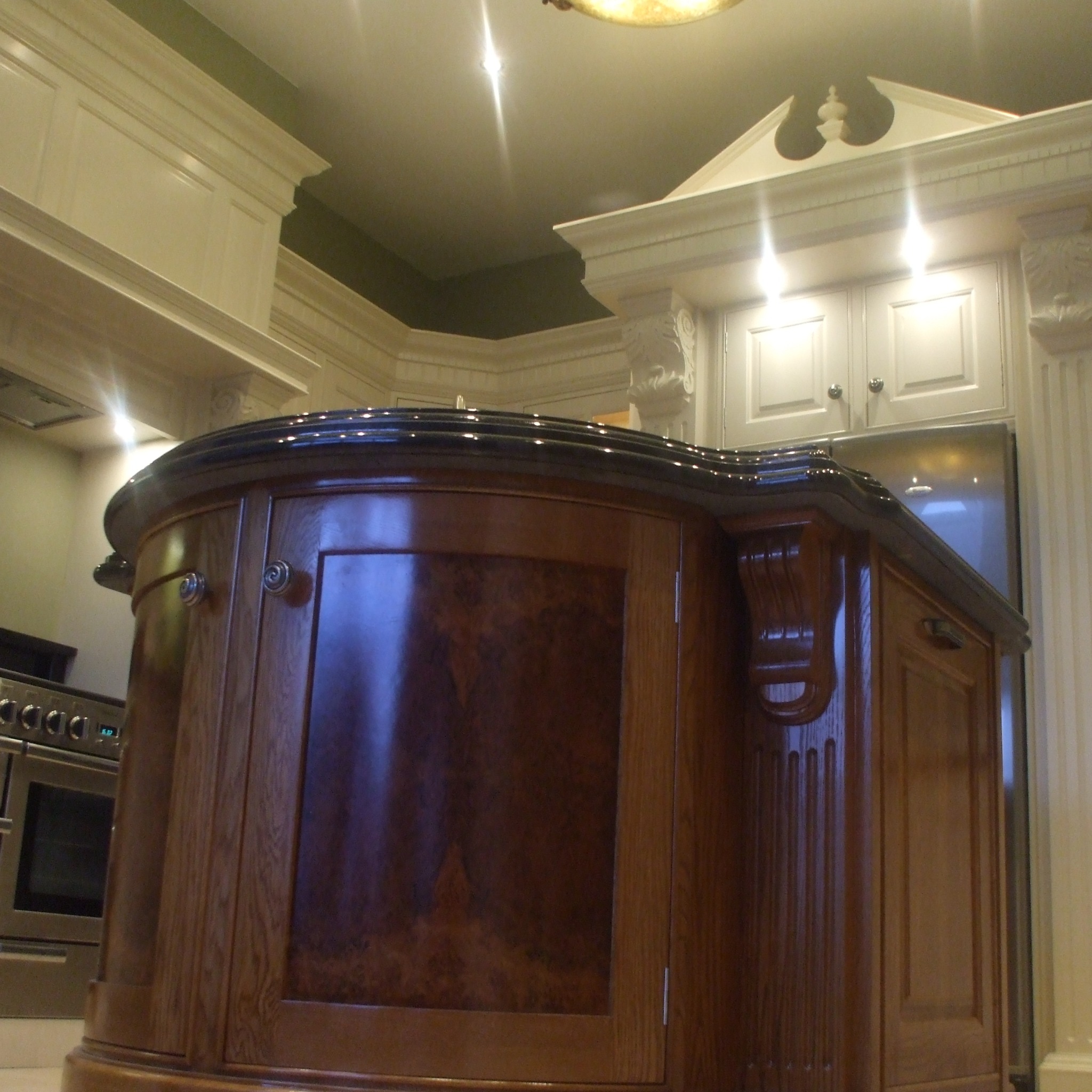 Zaokrąglona wyspa kuchenna z ciemnym blatem i ozdobnymi elementami, widok z dołu, w tle zabudowa kuchenna z białymi szafkami.