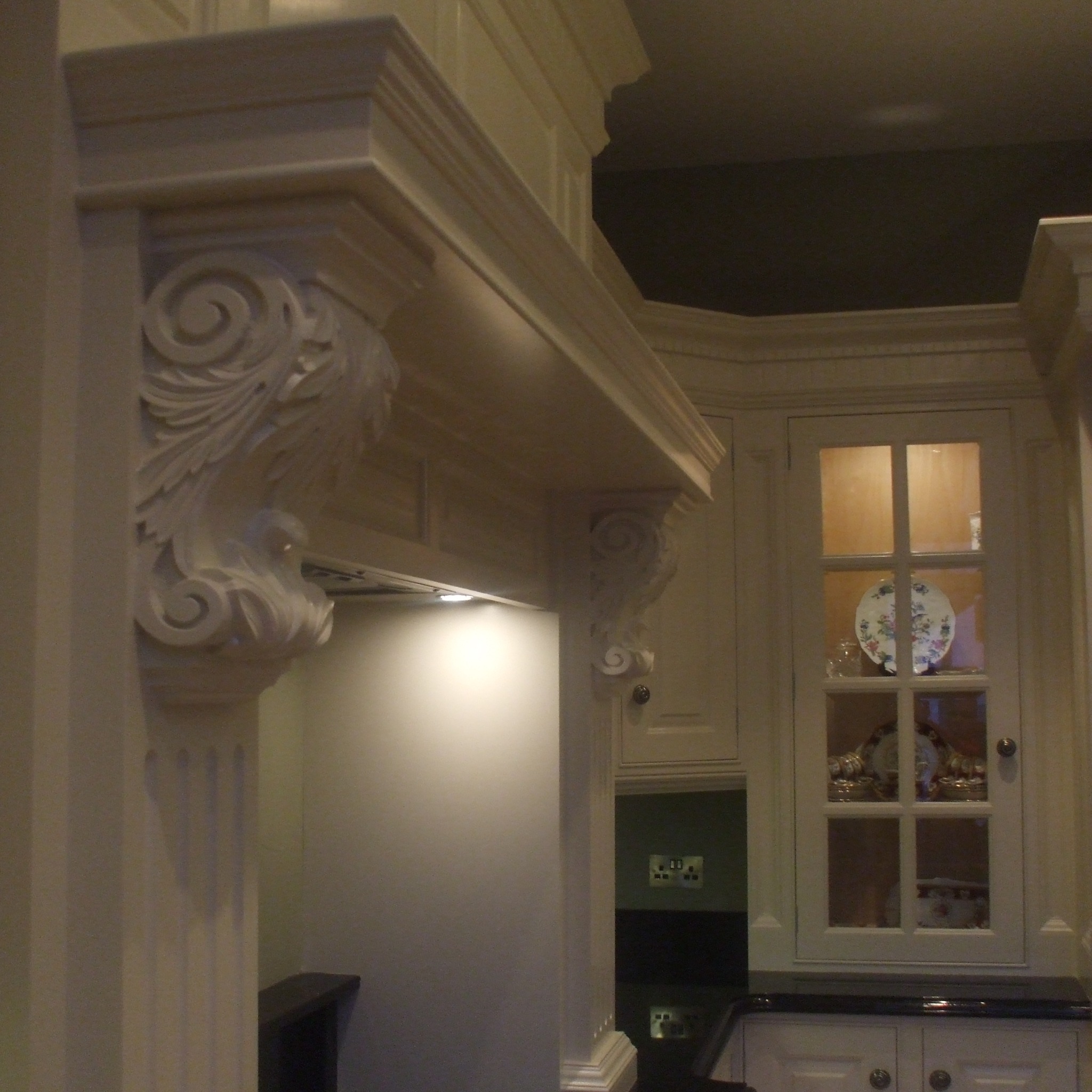 Fragment kuchni z ozdobnym okapem nad płytą grzewczą, białe szafki z przeszklonymi drzwiczkami i dekoracyjną porcelaną wewnątrz.