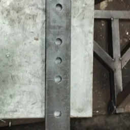 Metalowy element z otworami i spawami, leżący na stole warsztatowym, widoczne ślady obr&oacute;bki i rdzy.