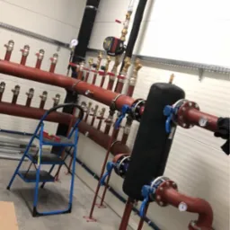 Rozbudowana instalacja grzewcza z czerwonymi rurami, zaworami i manometrami, widoczna drabina i skrzynka narzędziowa DeWalt na surowym betonowym podłożu w pomieszczeniu o ścianach z białej blachy...