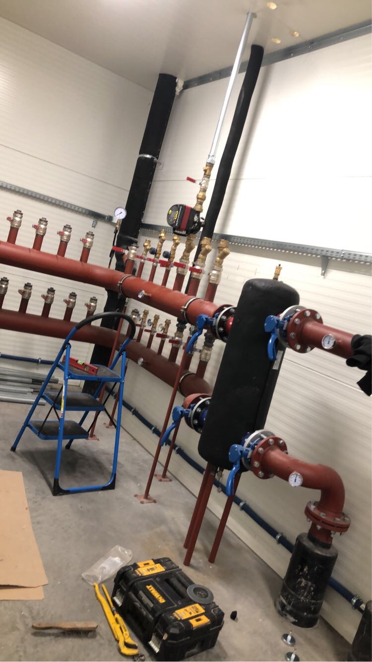 Rozbudowana instalacja grzewcza z czerwonymi rurami, zaworami i manometrami, widoczna drabina i skrzynka narzędziowa DeWalt na surowym betonowym podłożu w pomieszczeniu o ścianach z białej blachy...