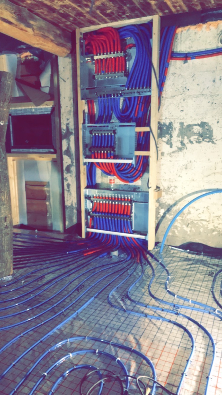 Rozdzielacz ogrzewania podłogowego z czerwono-niebieskimi rurami, zamontowany na ścianie w surowym pomieszczeniu, z widocznymi pętlami rur na metalowej siatce w trakcie instalacji.