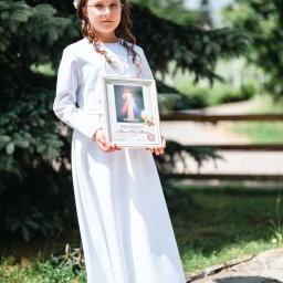 Natalia Kożuch fotografia - Dziewczynka w białej albie i wianku trzyma w rękach pamiątkowy obrazek z Pierwszej Komunii Świętej, na tle zieleni.