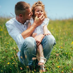 Natalia Kożuch fotografia - Tata przykucnął na łące pełnej kwiatów, trzymając na kolanach córeczkę, delikatnie ujmując jej twarz dłońmi. Dziecko ma kręcone włosy i srebrne sandałki.
