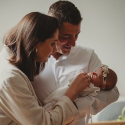 Natalia Kożuch fotografia - Rodzice z czułością spoglądają na noworodka z kwiatową opaską na głowie, ubranego w białą, koronkową sukienkę.