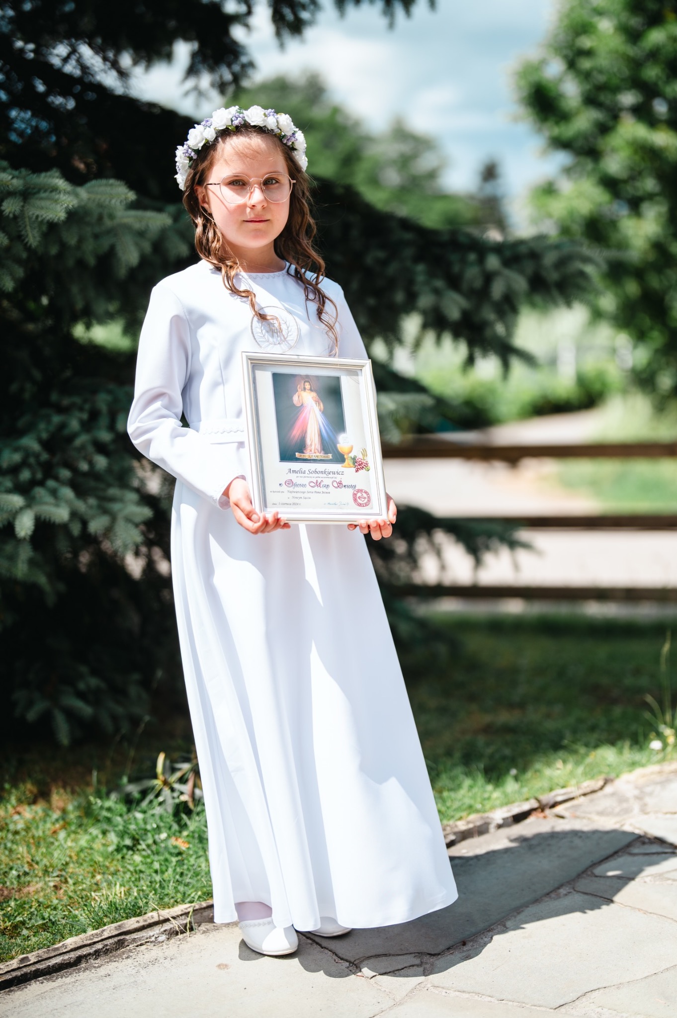 Dziewczynka w białej albie i wianku trzyma w rękach pamiątkowy obrazek z Pierwszej Komunii Świętej, na tle zieleni.