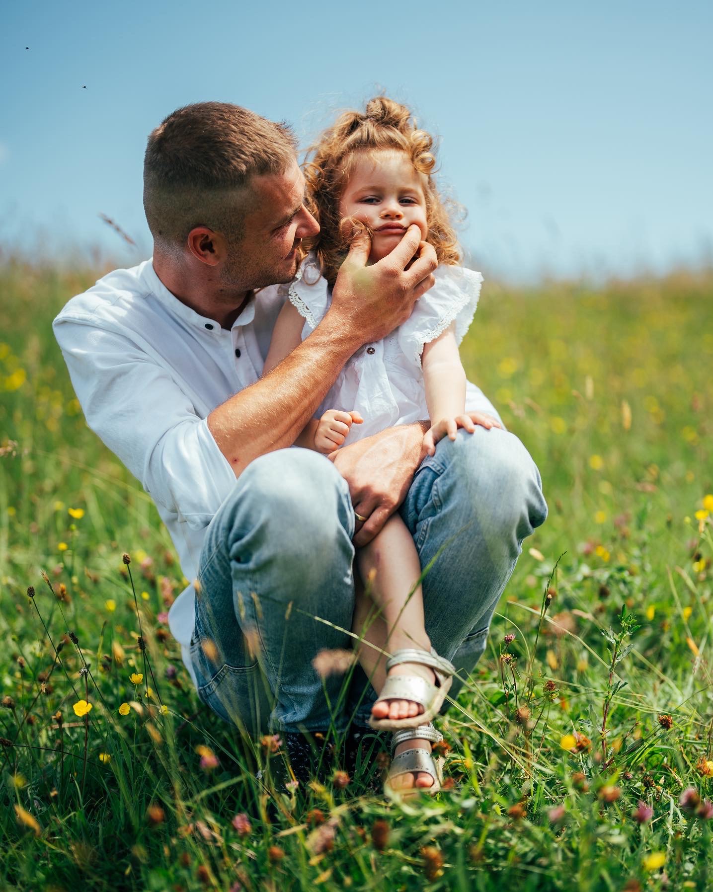 Tata przykucnął na łące pełnej kwiatów, trzymając na kolanach córeczkę, delikatnie ujmując jej twarz dłońmi. Dziecko ma kręcone włosy i srebrne sandałki.