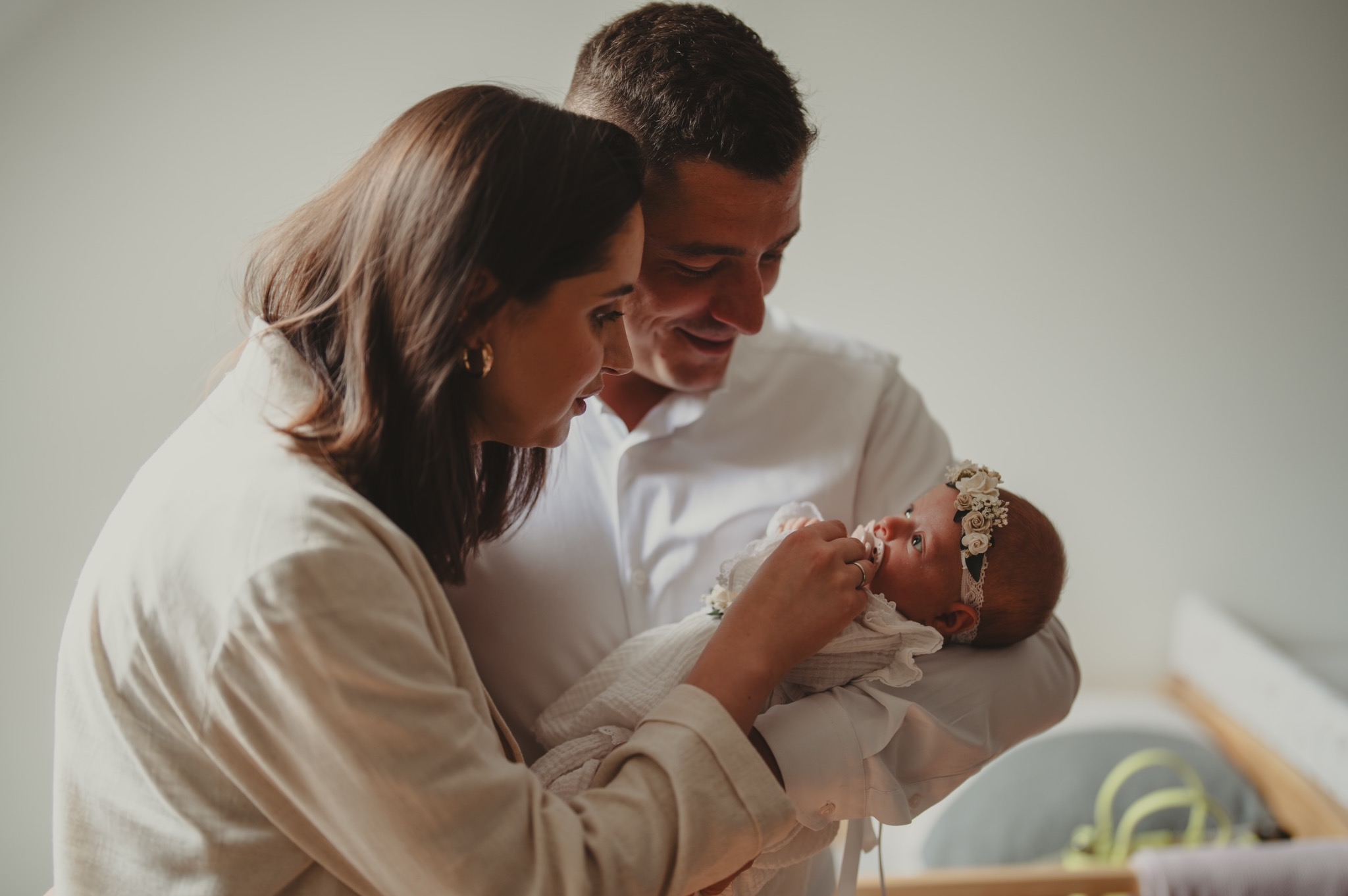 Rodzice z czułością spoglądają na noworodka z kwiatową opaską na głowie, ubranego w białą, koronkową sukienkę.