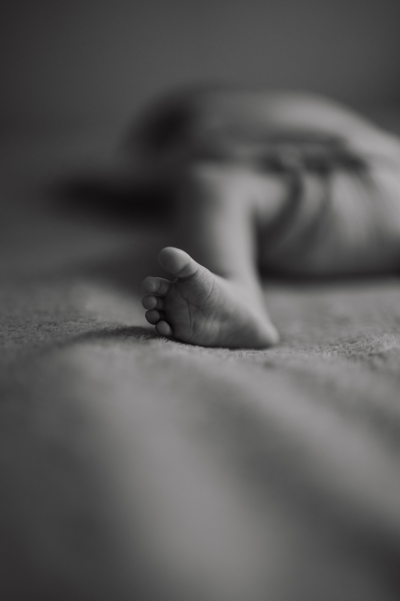 Mała stopa noworodka, delikatne paluszki na miękkim tle w czerni i bieli, głębia ostrości skupiona na szczegółach skóry.